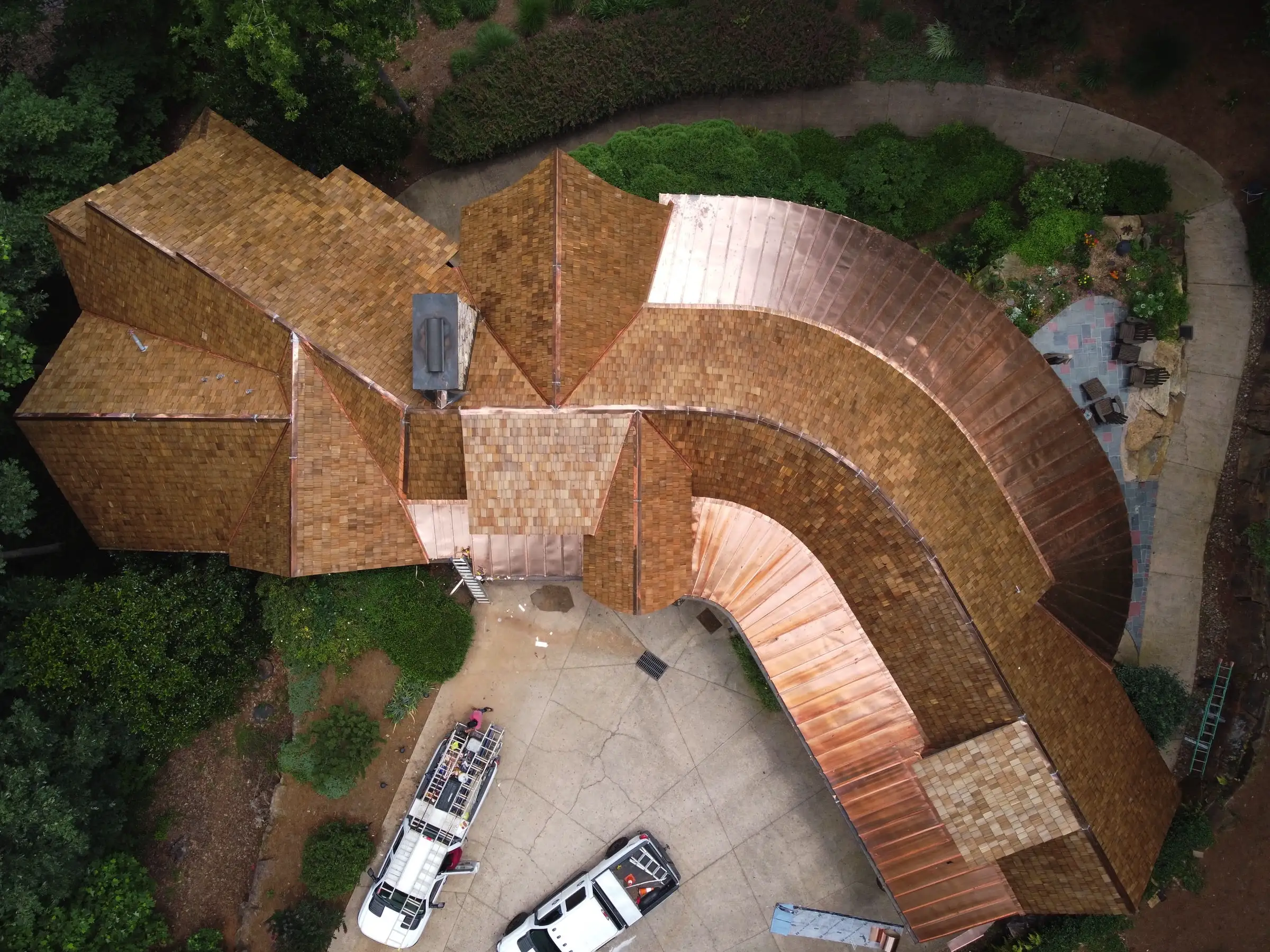 Aerial view of a complex residential roofing project featuring wood shake shingles and curved copper roofing sections, with work trucks and ladders visible in the driveway below.