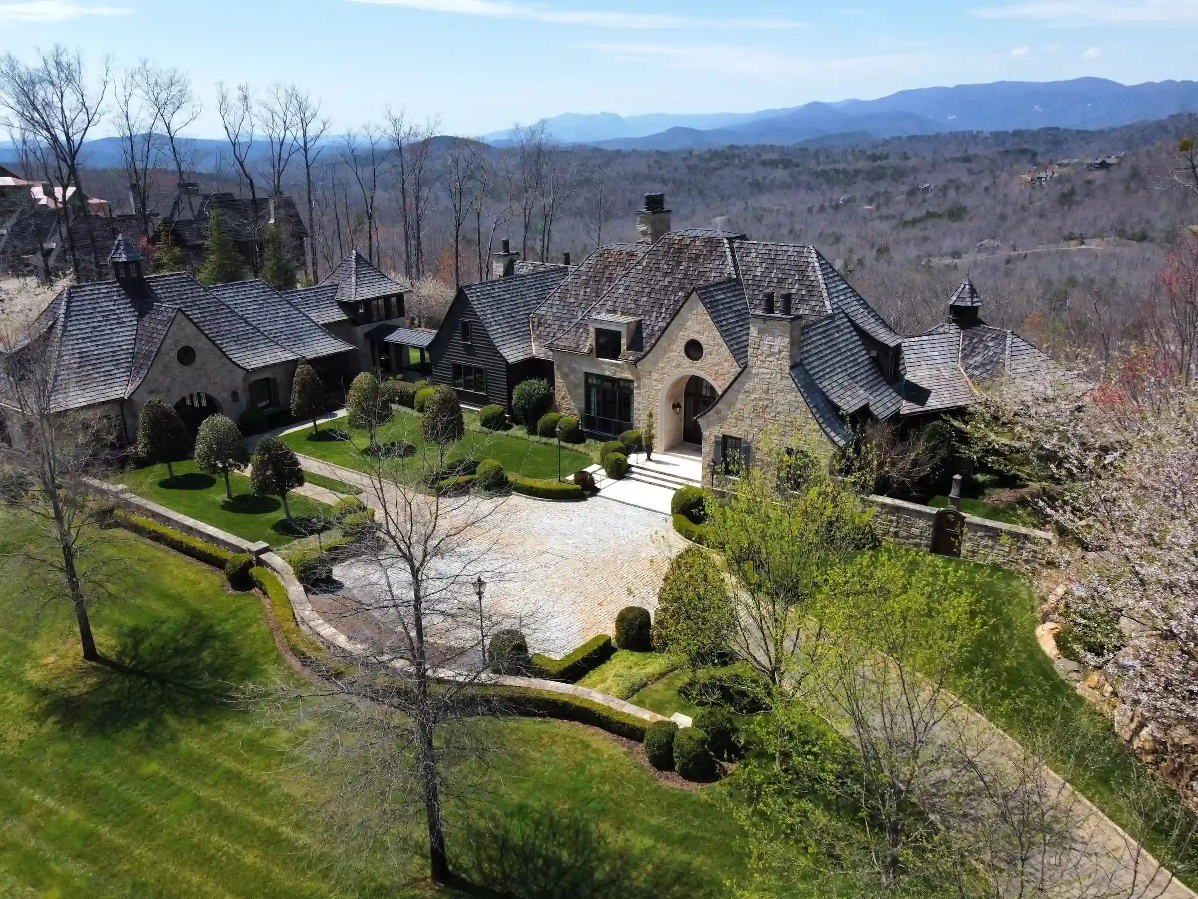 Aerial view of a large estate home with cedar shake roofing and stone exterior, surrounded by manicured landscaping and set against a backdrop of mountains and wooded hills.
